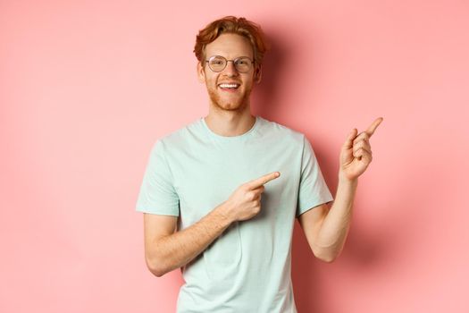 Friendly redhead guy in glasses pointing at upper right corner, showing promo banner and smiling at camera, standing over pink background.