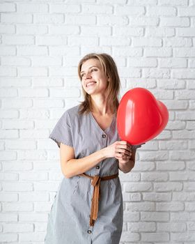 Valentines Day. Young woman holding read heart shaped balloon on white brick wall background