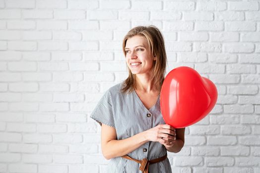 Valentines Day. Young woman holding read heart shaped balloon on white brick wall background with copy space
