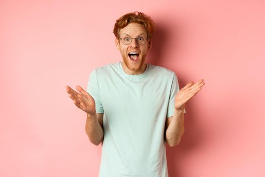 Image of cheerful young bearded man with red hair, wearing glasses, rejoicing of great news, spread hands sideways and congratulating you, standing over pink background.