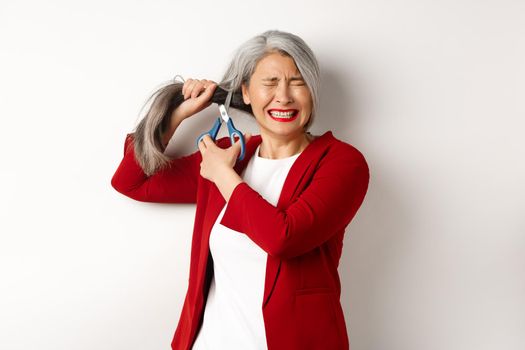 Scared asian woman cutting hair with scissors and feeling anxious, close eyes nervously, standing in red blazer against white background.