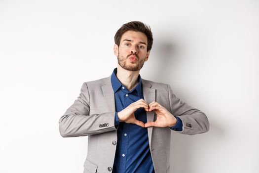 Romantic man in suit showing heart sign and pucker lips for kiss, express love and passion, like his lover, standing on white background.