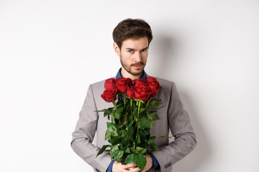 Handsome macho man going on date in suit, holding bouquet of red roses and smiling at camera, making valentines day surprise gift, white background.