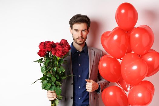 Valentines day. Handsome and confident man going on date in suit, holding bouquet of red roses and heart balloons, standing with gifts for lover on white background.