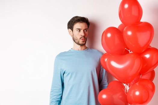 Relationship. Young man looking confused at heart balloon, puzzled on valetines day, standing over white background.
