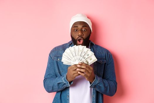 Amazed african-american man receive cash prize, showing money and staring in awe, standing over pink background. Copy space