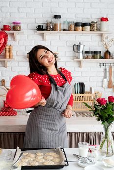 Valentines Day. Woman in red dress and gray apron making valentine cookies at the kitchen holding red heart balloon