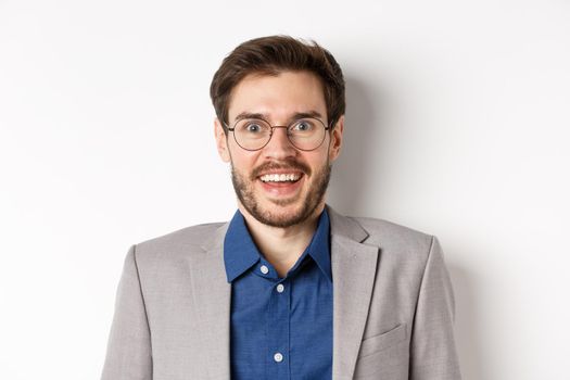 Cheerful european man in glasses and suit smiling excited, looking at camera with amazed happy face, white background.