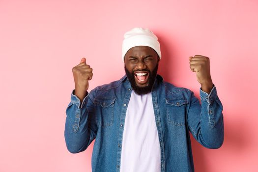 Image of happy Black man winning in lottery, rejoicing and making fist pump, scream yes in joy, standing over pink background.