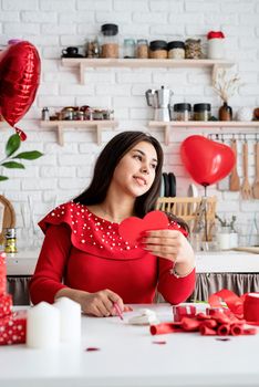 Valentines Day. Young romantic woman in red dress writing love letter sitting at the decorated kitchen