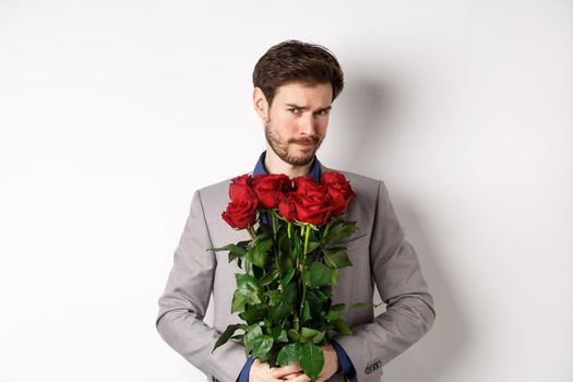Indecisive bearded man in suit, holding flowers roses and looking at camera unsure, going on romantic date, standing over white background.