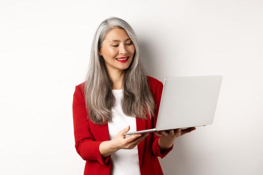Business. Successful asian businesswoman in red blazer working on laptop, smiling and reading screen, standing over white background.