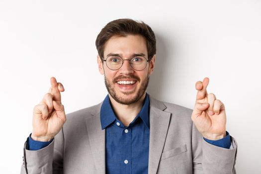 Close-up of hopeful male entrepreneur in glasses and suit cross fingers for good luck, smiling and making wish, praying for good results, white background.
