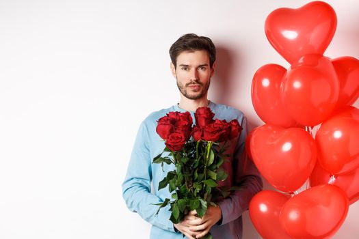 Valentines day romance. Confident young man holding bouquet of red roses, standing near hearts balloons, going on romantic date with lover, white background.