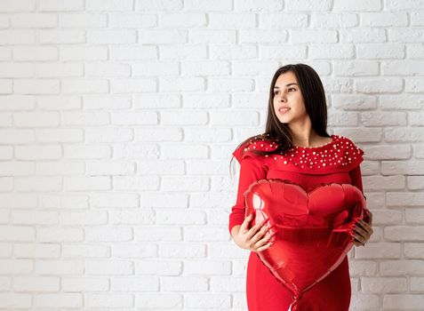Valentines Day. Young brunette woman in red dress holding a red heart balloon over white brick wall background