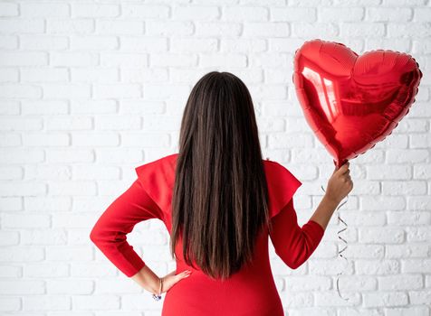 Valentines Day. Young brunette woman in red dress holding a red heart balloon over white brick wall background, view from behind