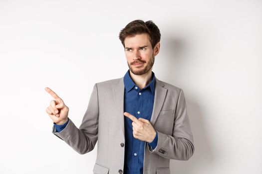 Displeased businessman in suit pointing and looking left with skeptical smirk, dislike banner, disapprove promo offer, standing on white background.