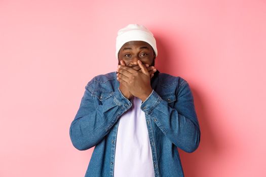 Happy Black hipster man holding laugh, cover mouth and giggle over funny joke, staring at camera and chuckling, standing over pink background.