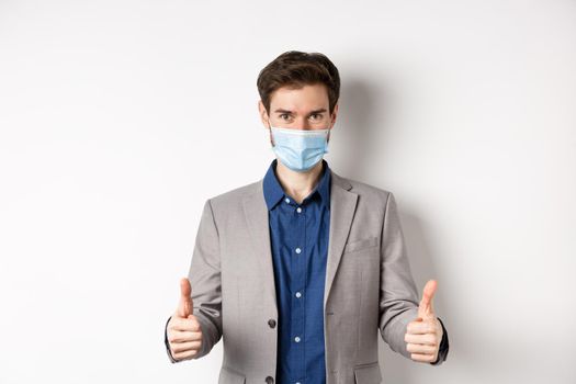 Covid-19, pandemic and business concept. Cheerful man in suit and medical mask using preventive measures in office, showing thumbs up, white background.