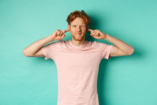 Annoyed young man with red hair and beard shut his ears and grimacing, disturbed by loud bothering sound, noisy neighbours, standing over turquoise background.