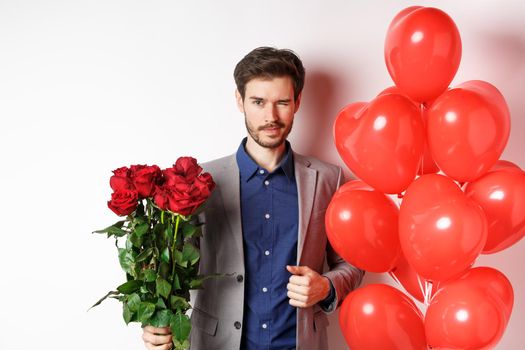 Confident boyfriend in suit going on romantic date, winking at camera, holding bouquet of red roses, standing near heart balloon and winking, standing over white background.