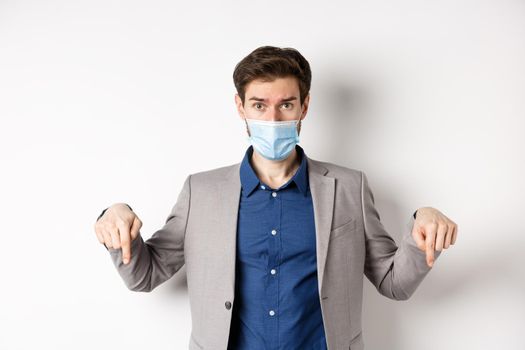Covid-19, pandemic and business concept. Hesitant man in suit and medical mask asking opinion, pointing fingers down and look at camera, white background.