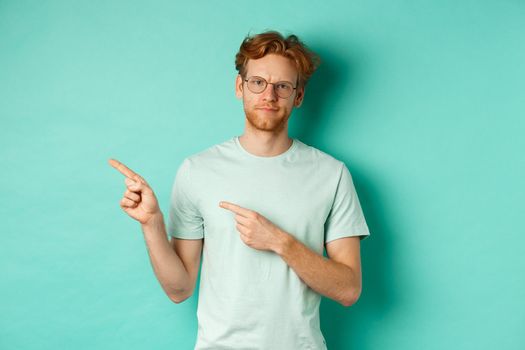 Judgemental redhead man in glasses grimacing, frowning disappointed and pointing left, showing bad promo offer, standing over turquoise background.