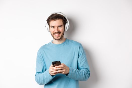 Attractive smiling man listening music in wireless headphones, using black smartphone and looking pleased, white background.