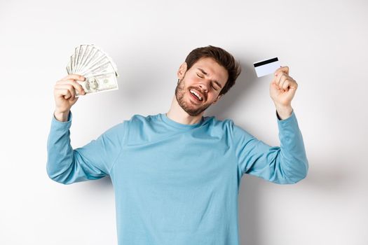 Handsome man dancing with money and plastic credit card, standing in casual clothes over white background.