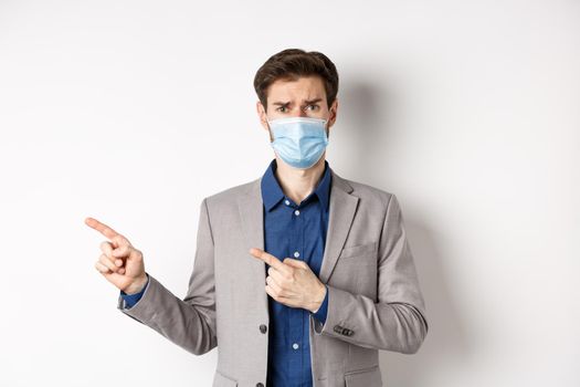 Covid-19, pandemic and business concept. Upset man in medical mask and office suit, frowning sad and pointing left at banner, standing on white background.