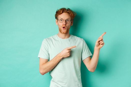 Surprised redhead guy in glasses and t-shirt checking out special deal, pointing at upper right corner promo and gasping in awe, showing banner, standing over turquoise background.