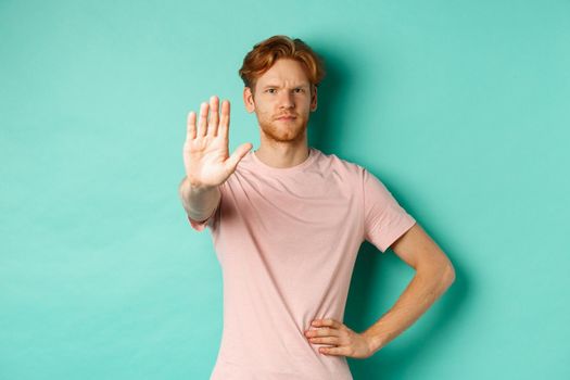 Serious young man with red hair and beard telling to stop, showing palm and frowning, disapprove and prohibit something bad, standing over turquoise background.