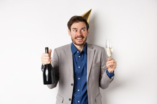 Celebration and holidays concept. Happy birthday guy in suit and party hat drinking champagne, holding bottle and glass, smiling and looking aside.