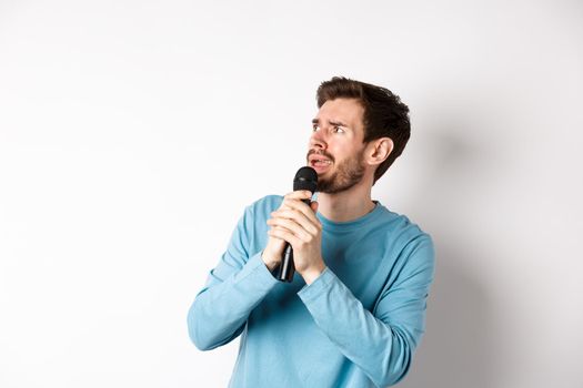 Worried and confused man reading lyrics on karaoke, looking left with unsure face, holding microphone and singing, white background.