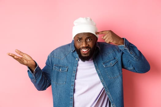 Annoyed and pissed-off african-american man pointing finger at head, scolding someone stupid, staring bothered at camera, pink background.