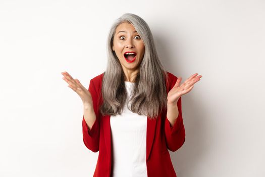 Business people. Surprised senior asian female entrepreneur, gasping amazed, staring at camera, hear great news, standing happy over white background.