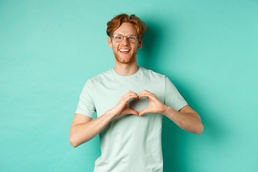 Valentines day and relationship concept. Happy boyfriend with red hair and beard, wearing glasses and t-shirt, showing heart sign and saying I love you, standing over turquoise background.