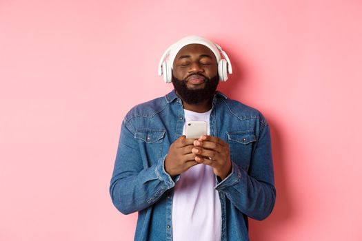 Delighted Black man enjoying awesome music, listening songs in headphones and holding smartphone, looking ecstatic, standing over pink background.