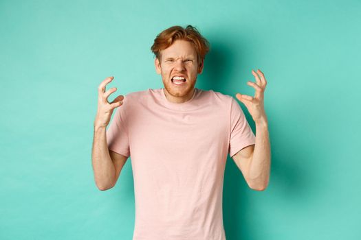 Annoyed and pissed-off redhead guy looking angry, shouting and grimacing, shaking hands aggressive, standing furious against mint background.