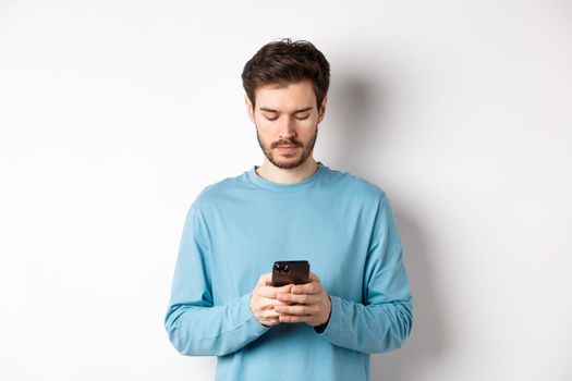 Serious young man reading message on smartphone, looking at mobile screen, standing over white background.