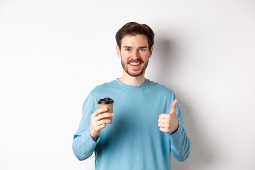 Cheerful young man drinking good coffee, holding paper cup and showing thumb up, recommending cafe shop, standing over white background.