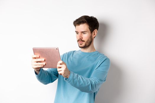 Young man taking photo on digital tablet, looking serious at gadget screen, standing over white background.