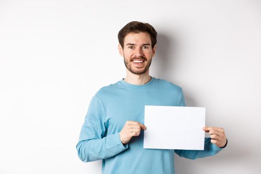 Young man with happy face showing blank piece of paper for your logo or sign, smiling at camera, standing over white background.