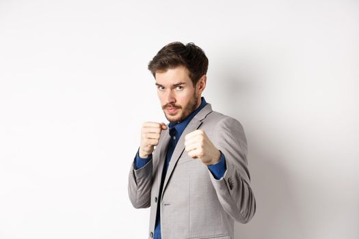 Serious man in suit raising hands in boxer pose, going to fight, looking confident, beckon to attack, standing on white background in defensive pose.