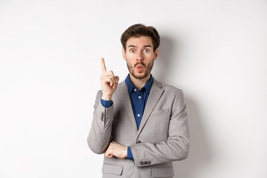 Excited businessman in suit pitching good idea, raising finger in eureka sign and looking amused, saying suggestion, standing on white background.