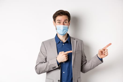 Covid-19, pandemic and business concept. Excited male entrepreneur in medical mask and suit pointing fingers right and looking curious, white background.