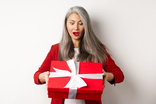 Surprised asian senior woman receiving present on mother day, holding red box with gift and looking amazed, white background.