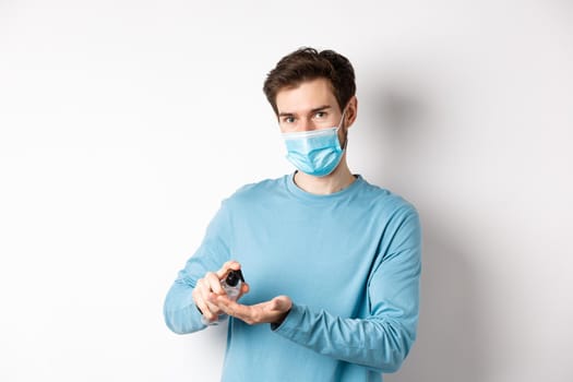 Covid-19, health and quarantine concept. Caucasian man in medical mask using hand sanitizer, apply antiseptic from coronavirus spread, white background.
