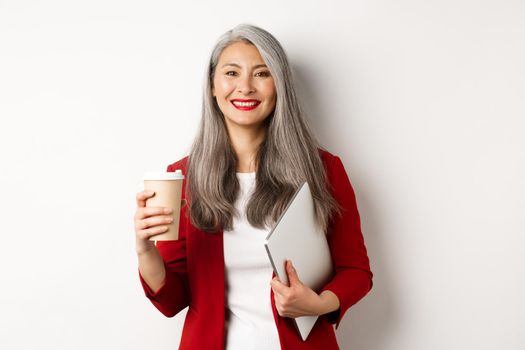 Business. Successful asian female manager standing with cup of coffee and laptop, smiling satisfied at camera, white background.