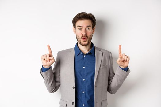 Excited businessman in suit say wow and smiling amused, pointing fingers up at good deal, standing against white background.